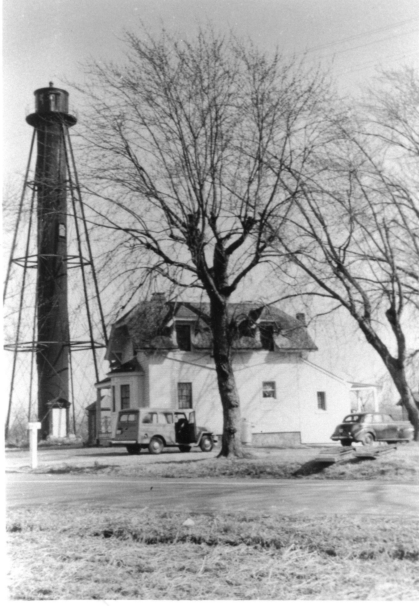 Vintage black-and-white photo of a tall skeletal tower beside a white house, with vintage vehicles parked nearby