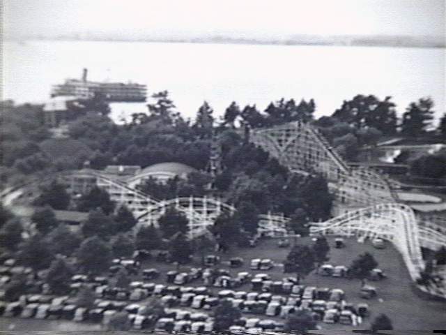 Black and white aerial view of an amusement park with roller coaster, trees, and parked cars near water