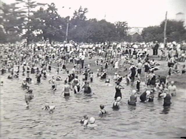 Black and white photo of a large crowd wading in shallow water at a beach, overcast mood